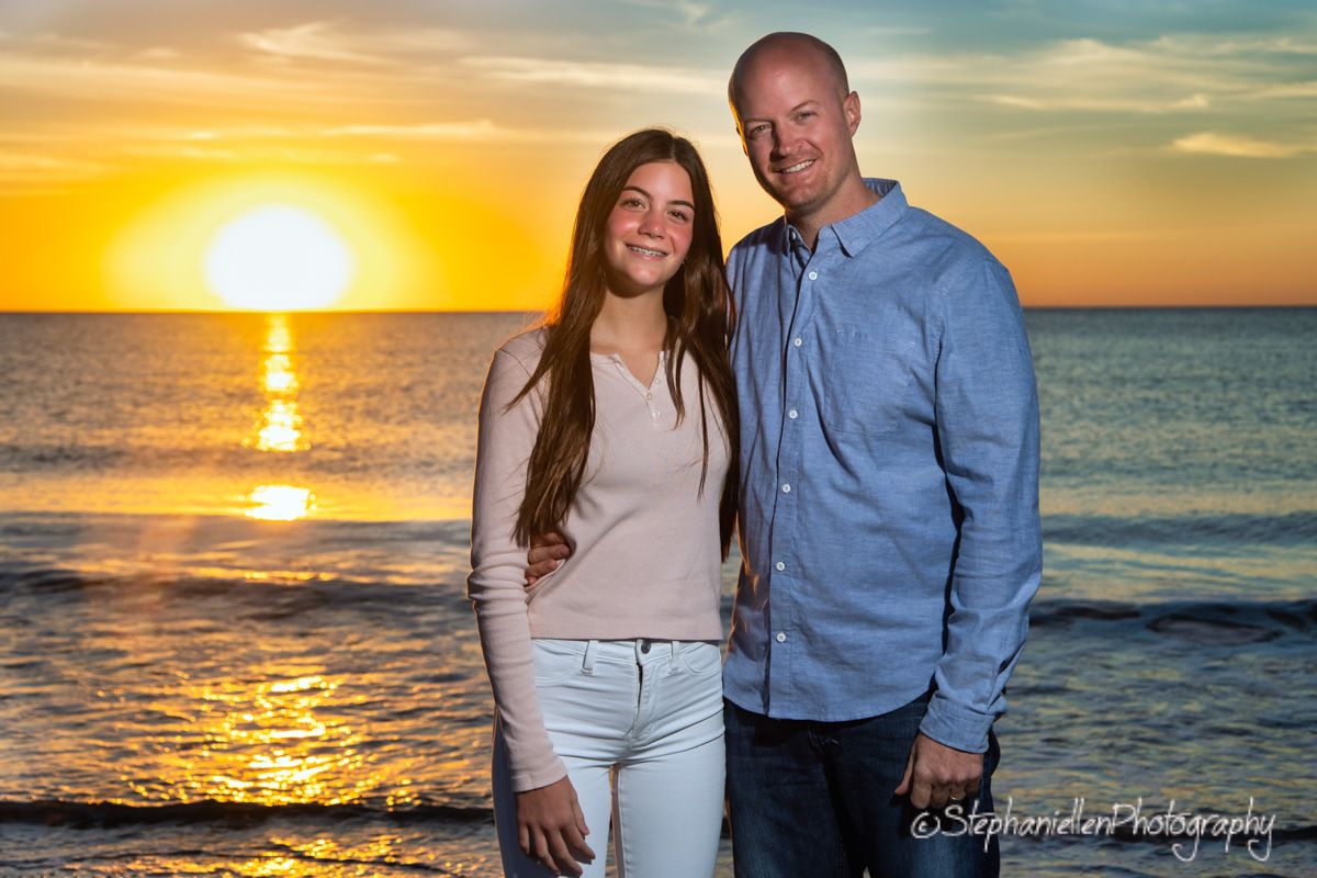 A couple smiles together on a beach at sunset with golden light reflecting off the ocean behind them.