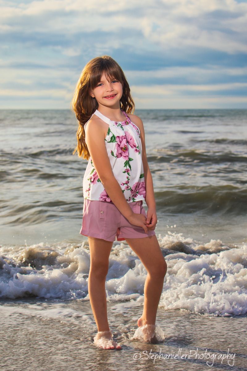 Young girl smiling at beach in floral top and pink shorts, waves splashing around her feet.