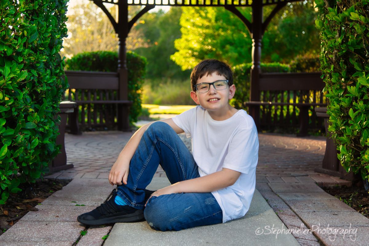 A smiling boy with glasses sits on a brick path in a lush green garden, wearing a white shirt and jeans.
