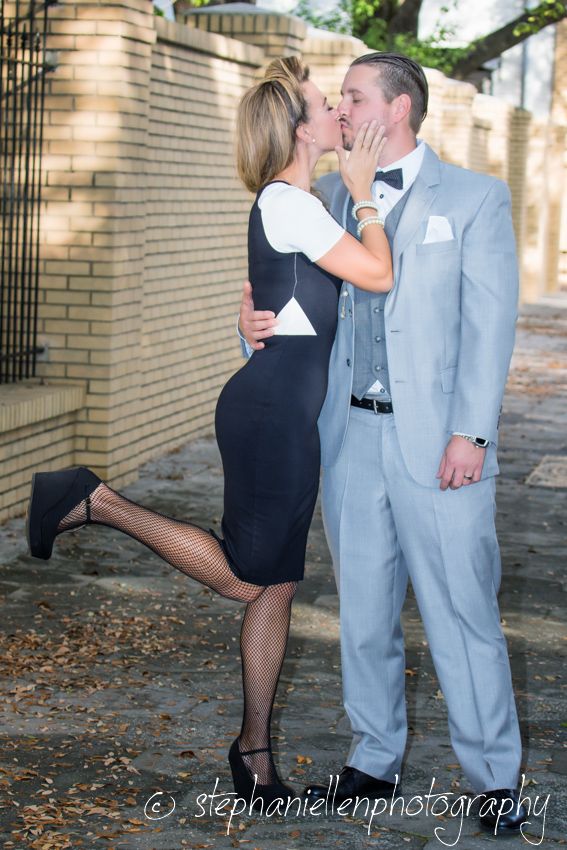 Couple sharing a romantic kiss outdoors, man in gray suit with bow tie, woman in black dress with fishnet stockings.