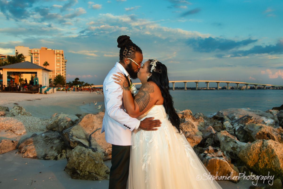 Newlyweds share romantic kiss on rocky beach at sunset with bridge in background.