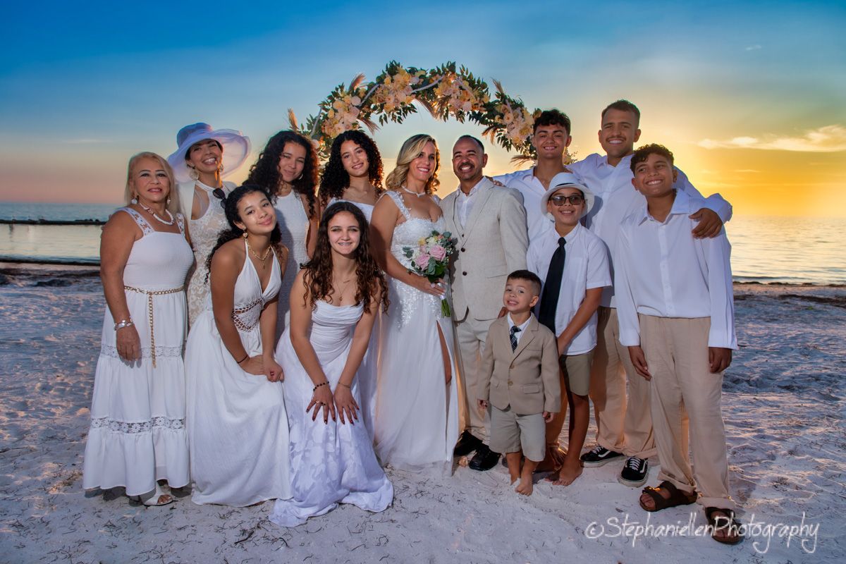 Wedding party group photo on the beach at sunset with floral arch backdrop.