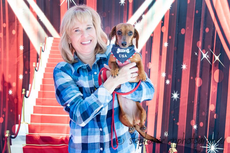 Woman in plaid shirt holds a dachshund wearing a USA vest, posing on a red carpet with stars backdrop.