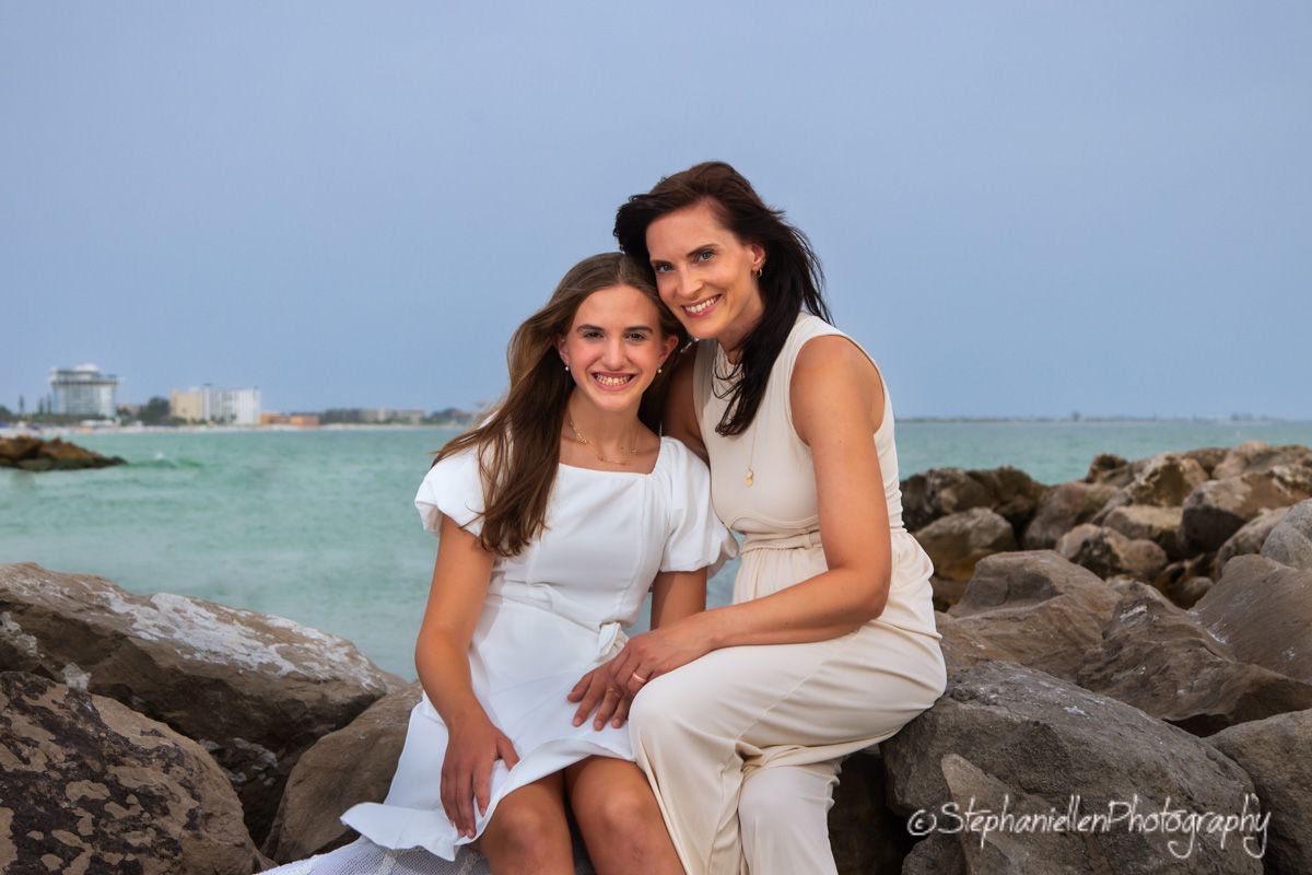Two women in white dresses smile together on rocky shoreline with turquoise ocean and blue sky backdrop.