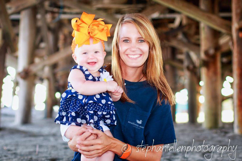 Smiling mother holding her baby girl in a polka dot dress under a pier at the beach.