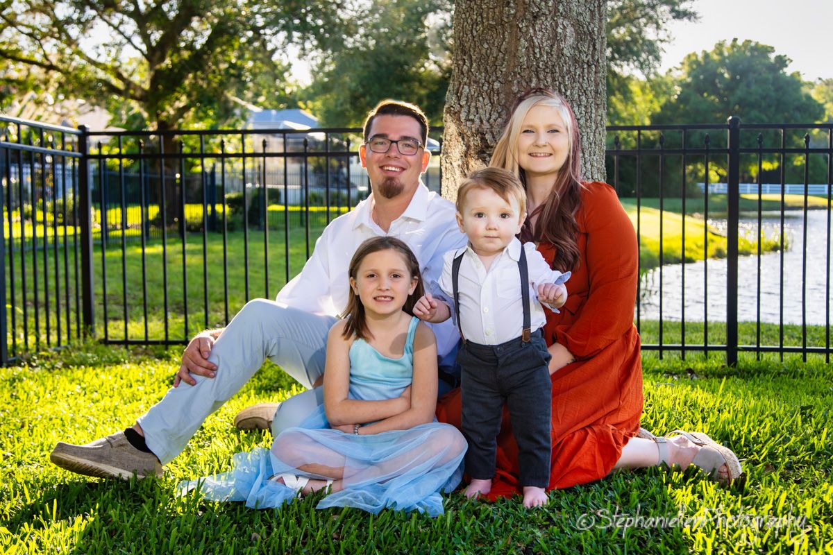 Family of four posing outdoors near a tree on green grass, smiling in coordinated outfits during golden hour.