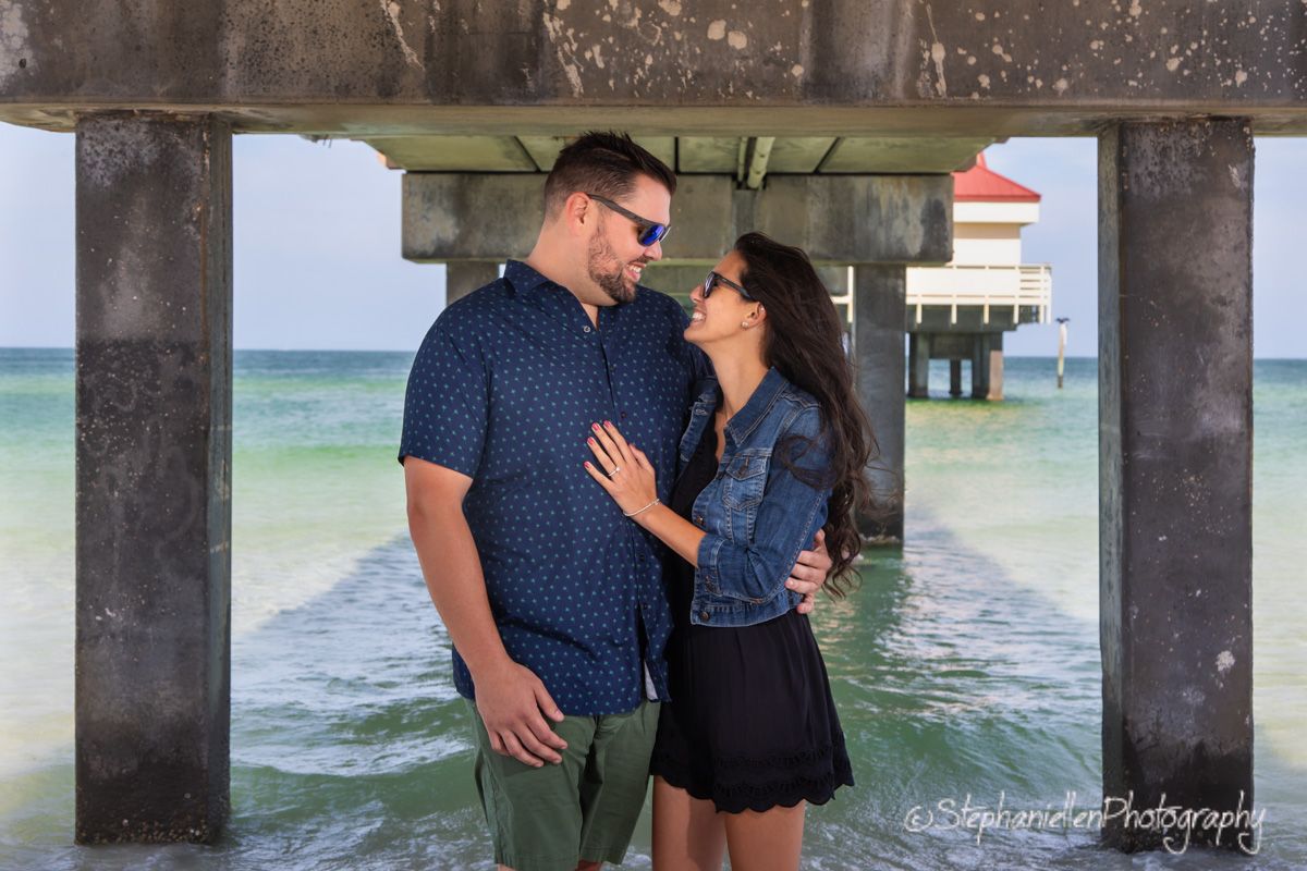 Happy couple embracing and smiling at each other under a pier with turquoise ocean behind them.