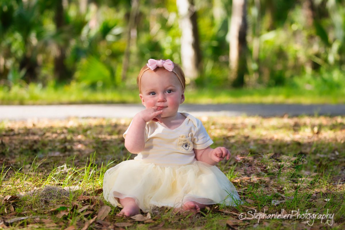 Adorable baby girl in yellow tutu dress sitting on grass with hand on chin in park.