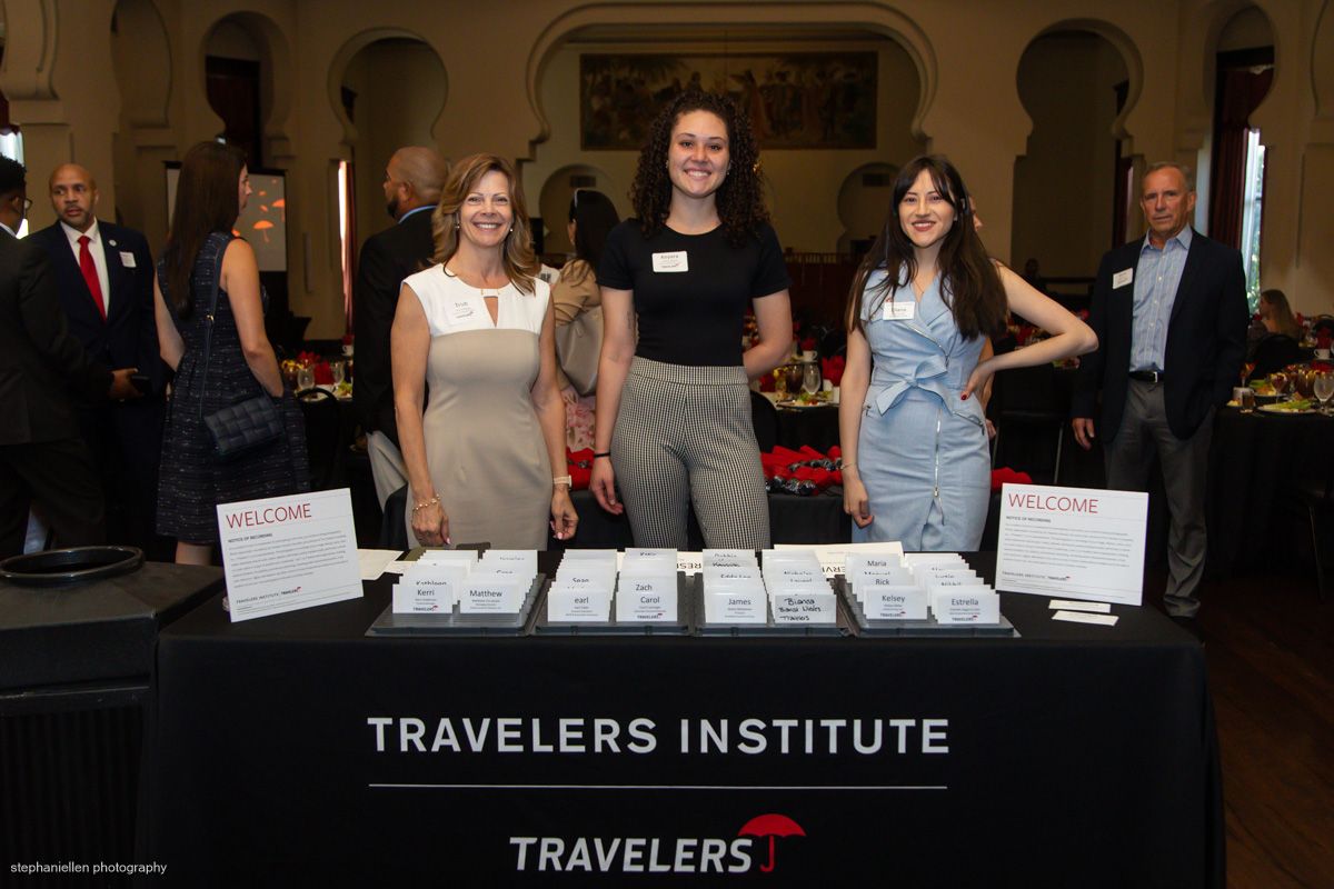 Three women stand behind a Travelers Institute welcome registration table at a formal event.