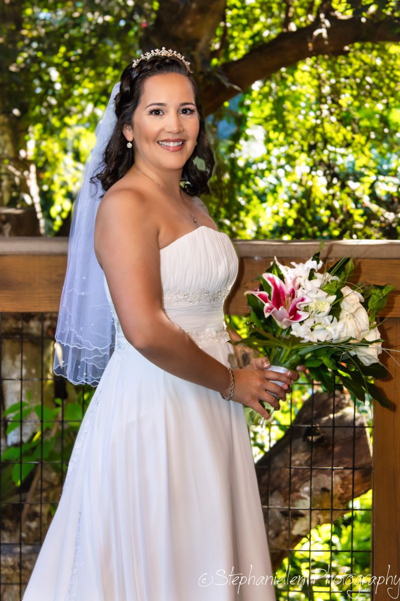 Smiling bride in white strapless gown holding a pink lily bouquet on a wooden bridge outdoors.