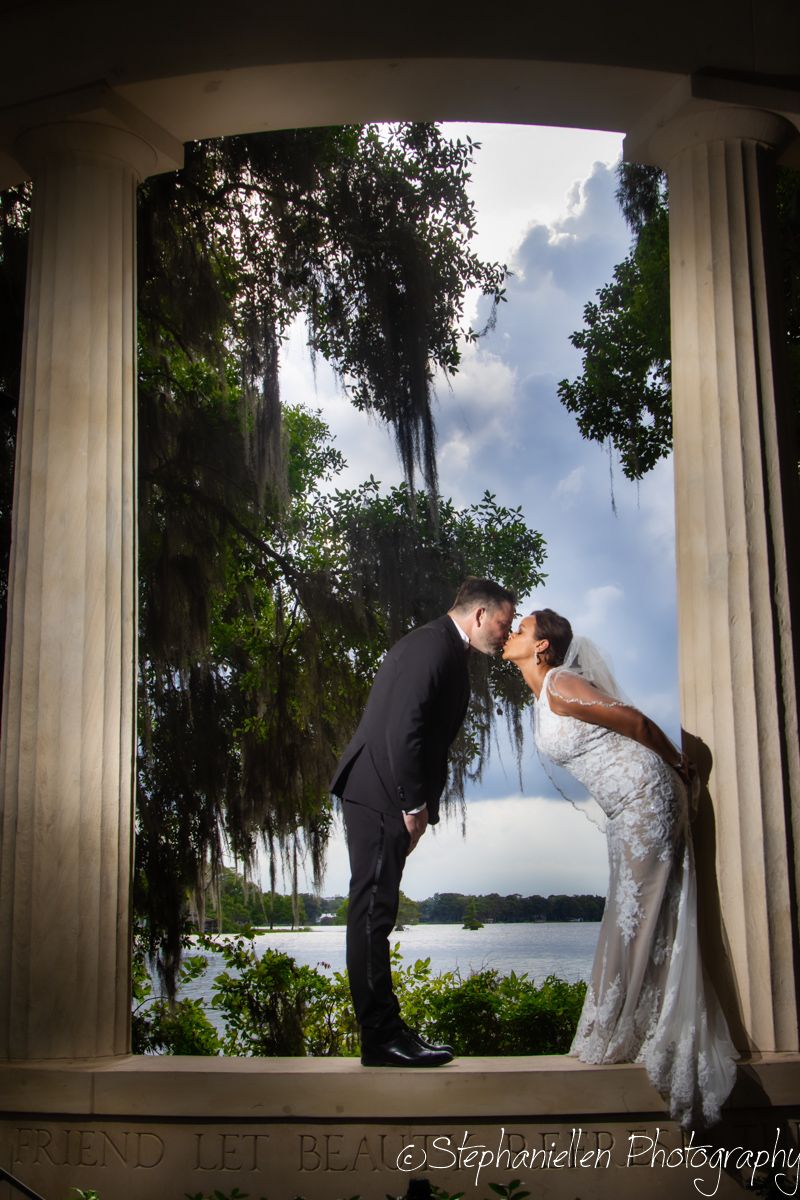 Bride and groom share romantic kiss framed by columns overlooking a serene lake with Spanish moss-draped trees.
