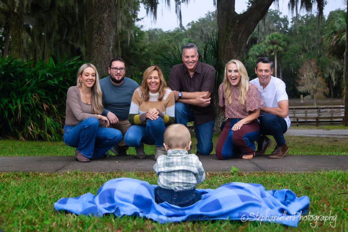 Family of six poses outdoors with a baby sitting on a blue blanket in foreground.