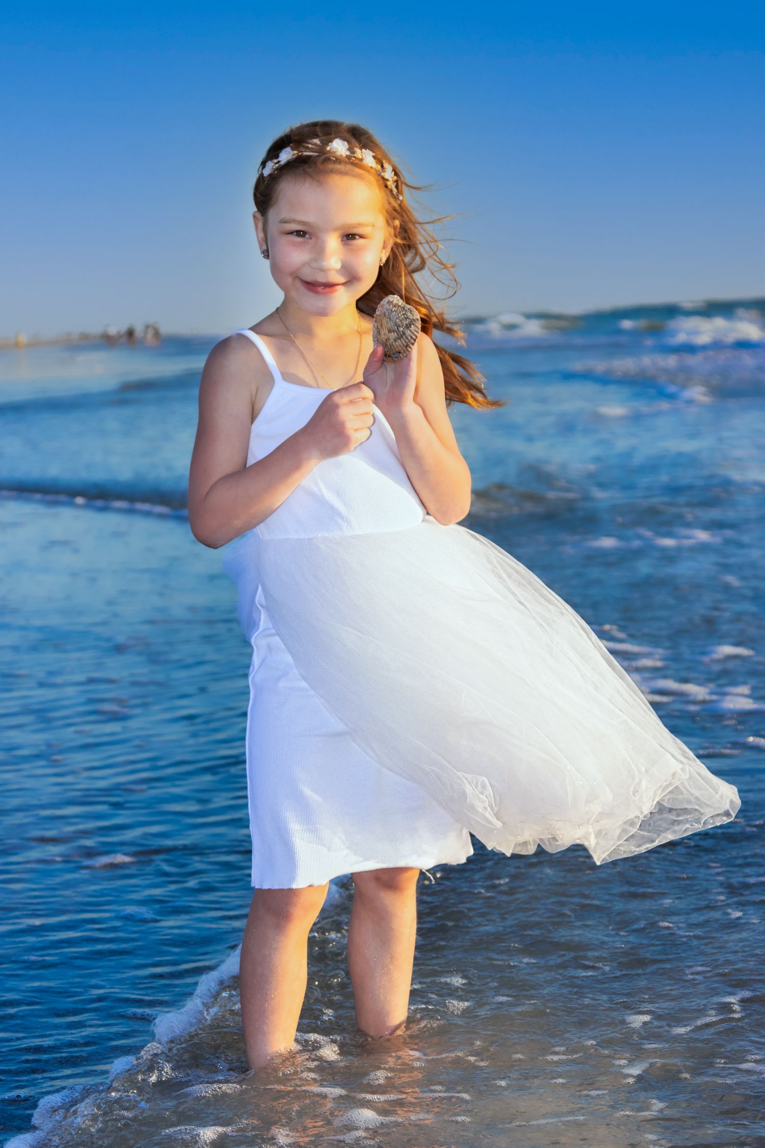 Young girl in white dress standing in ocean waves on a sunny beach, smiling and holding a seashell.