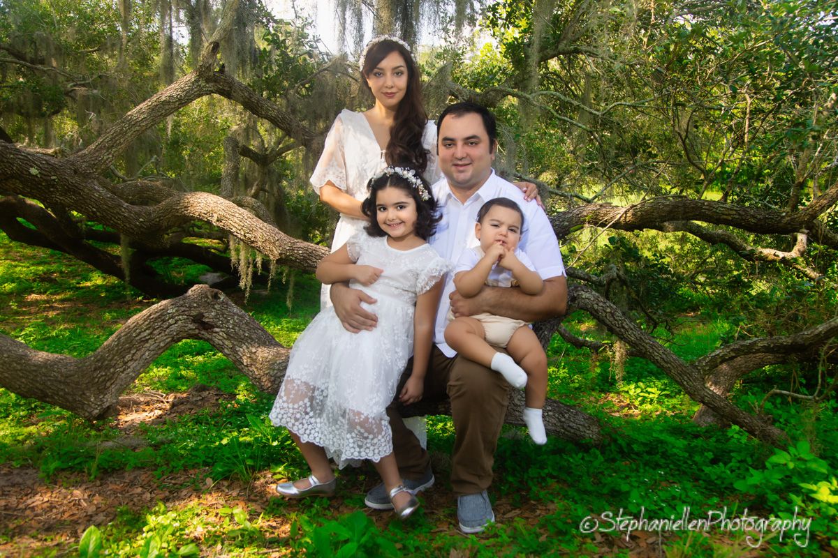 Family of four posing outdoors among mossy trees, parents with two young children in white outfits.