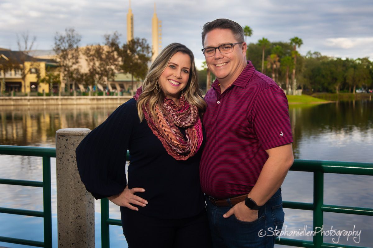A smiling couple poses together on a waterfront bridge at sunset with lush greenery and calm water in the background.