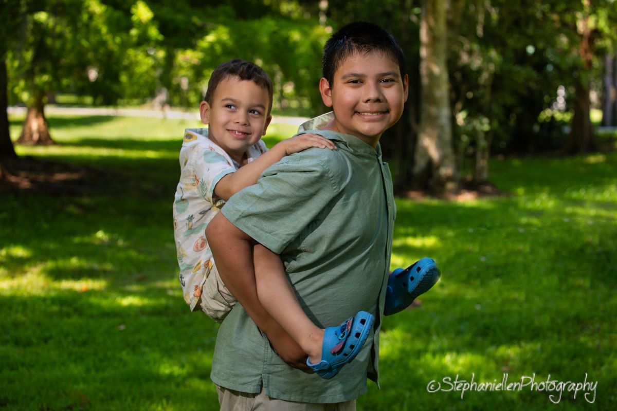 Two boys smile outdoors in a park, with the older boy giving the younger a piggyback ride on a sunny day.
