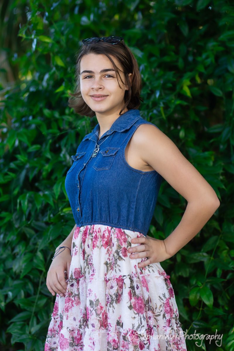 Young woman in denim top and floral skirt smiling outdoors against lush green foliage backdrop.