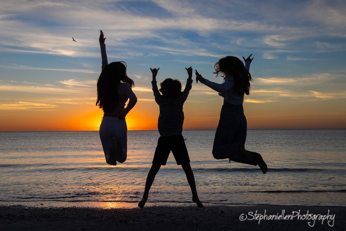 Three silhouettes jumping joyfully on a beach at sunset with vibrant orange and blue sky backdrop.