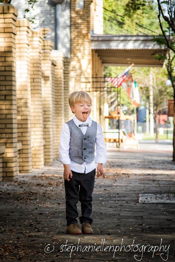 Adorable blonde toddler boy in gray vest and bow tie laughing outdoors near brick building.