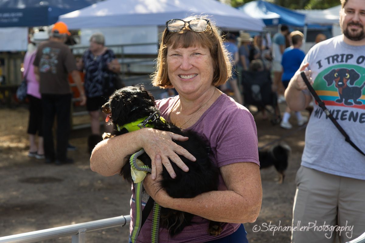 Happy woman smiling while holding a small black dachshund dog at an outdoor pet festival event.