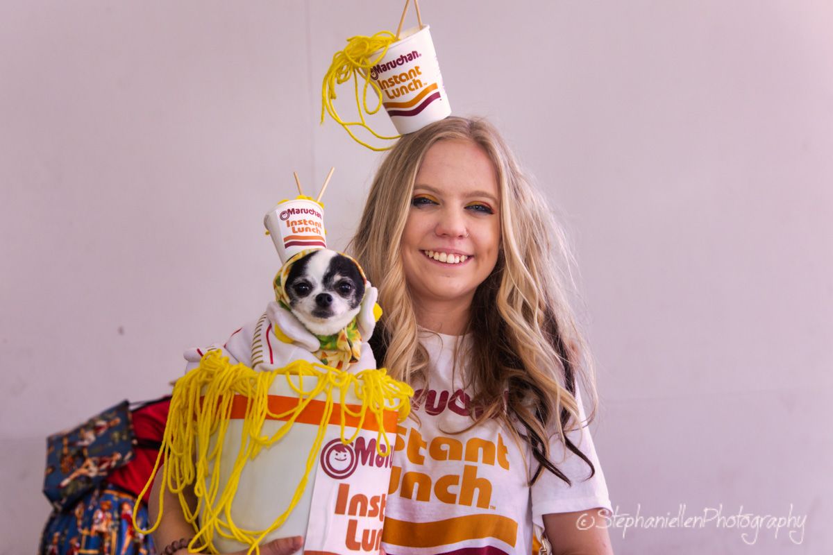 Smiling blonde girl in Instant Lunch costume holds small dog dressed in matching ramen noodle outfit for Halloween.