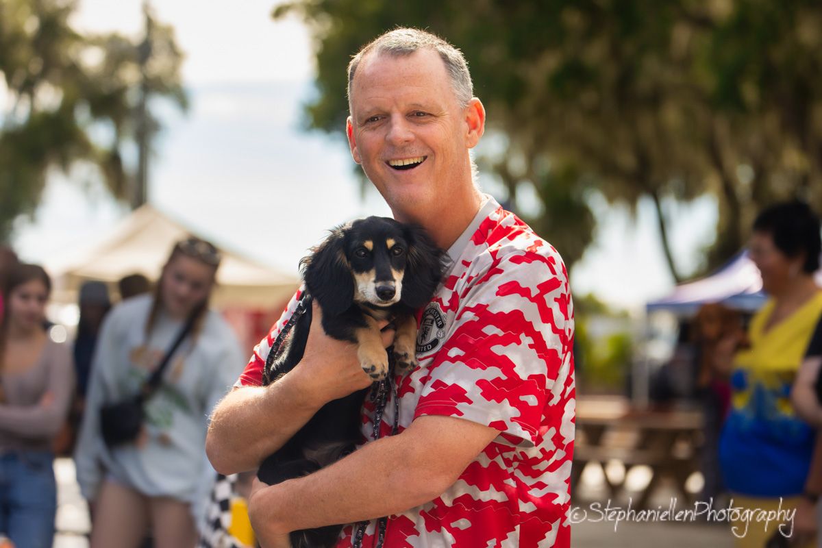 Smiling man in red floral shirt holding a small black and tan dog at an outdoor event.