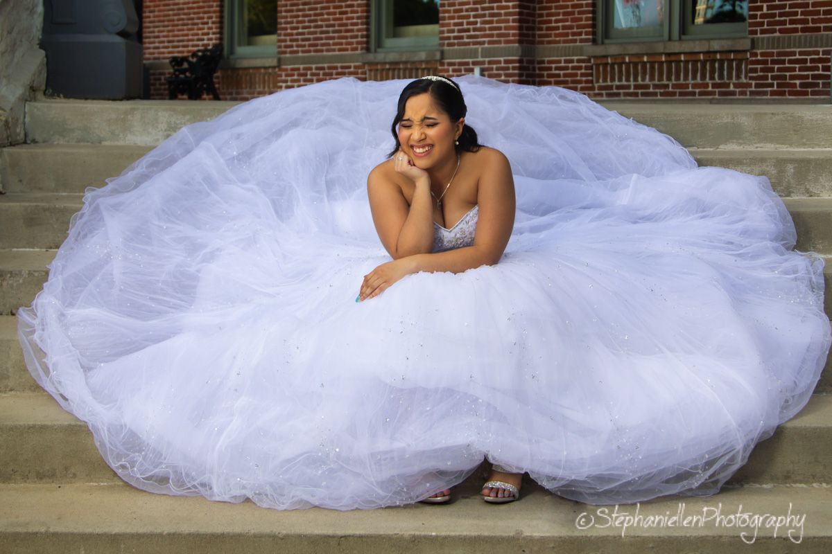 Bride in a voluminous white ball gown sitting gracefully on stone steps, smiling beautifully for wedding portrait.