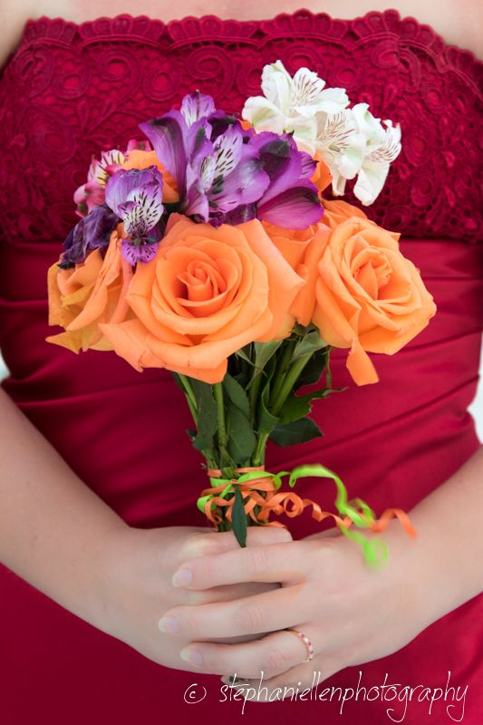 Bridesmaid in red dress holding a vibrant bouquet of orange roses and purple flowers at a wedding.