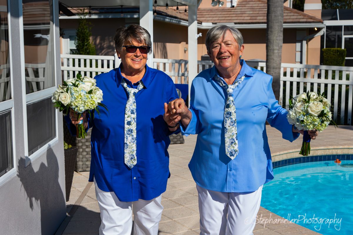 Two women in matching blue outfits and floral ties celebrate joyfully outdoors near a pool on their wedding day.