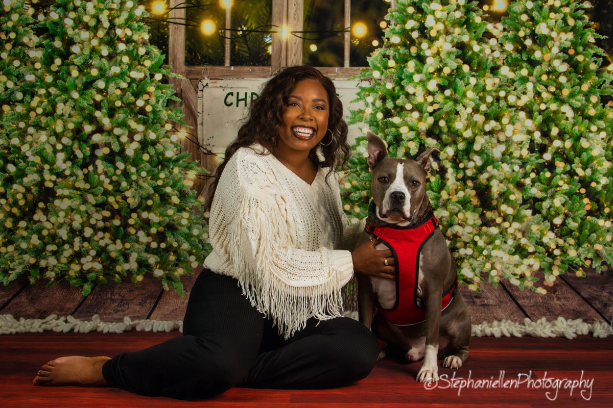 Woman smiling with blue pit bull in red harness against holiday floral light backdrop.