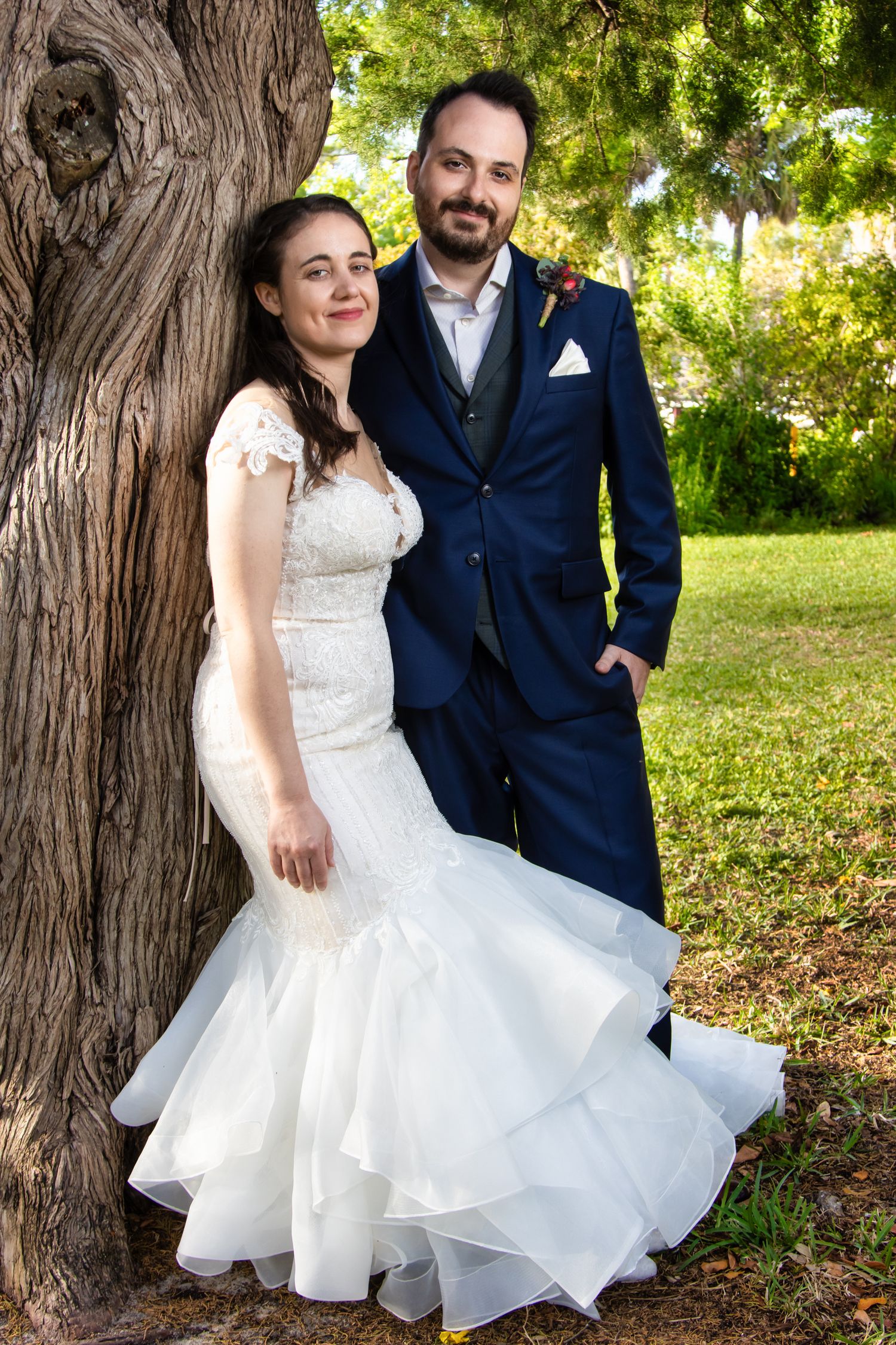 Bride in white mermaid gown and groom in navy suit pose together against a tree in a lush green park.