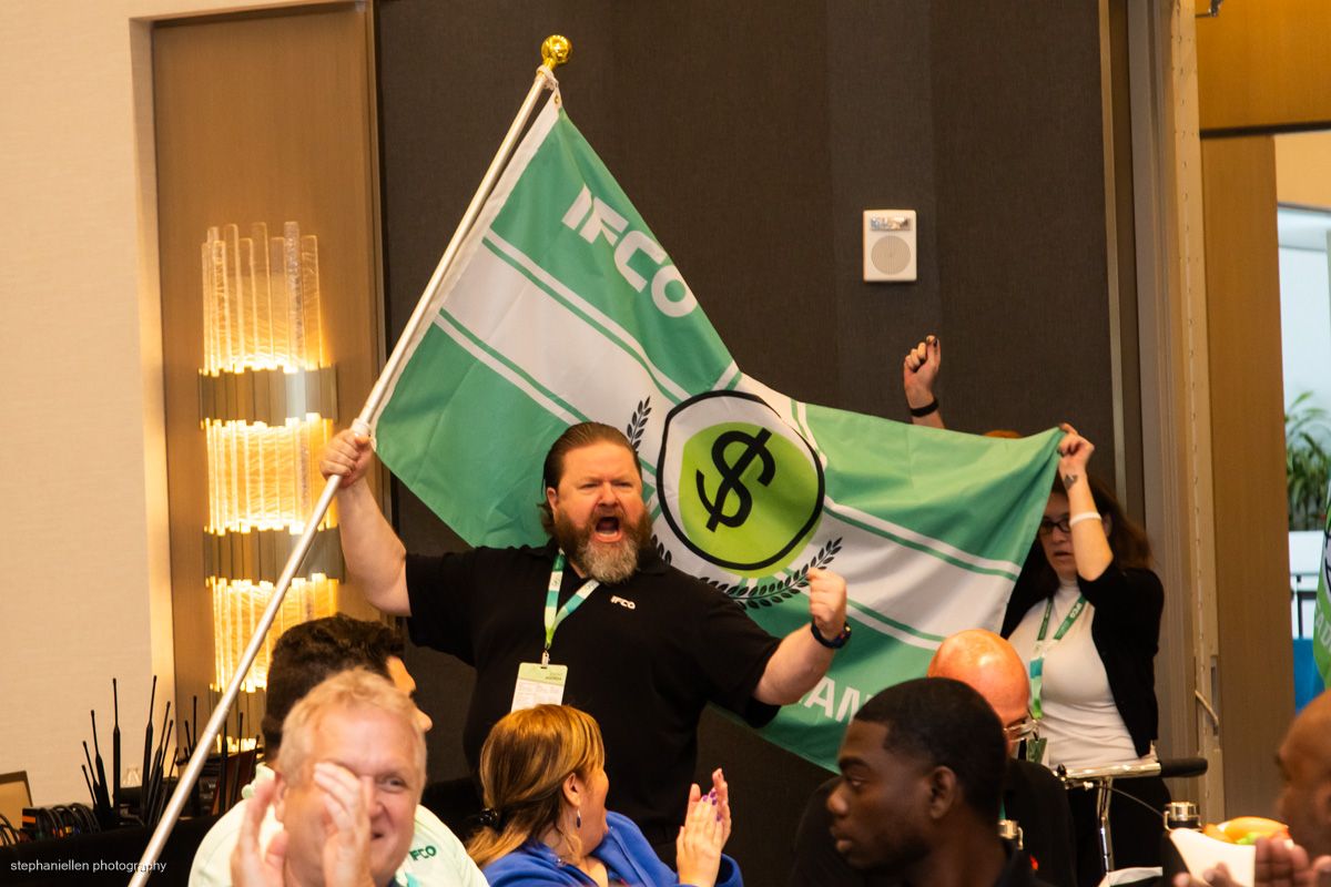 Enthusiastic man waves IFCO flag at an indoor conference or convention event.