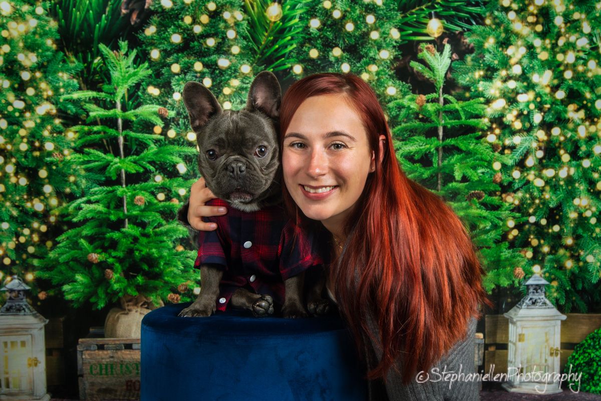 Smiling woman with red hair holding a French Bulldog puppy in plaid jacket among Christmas trees.