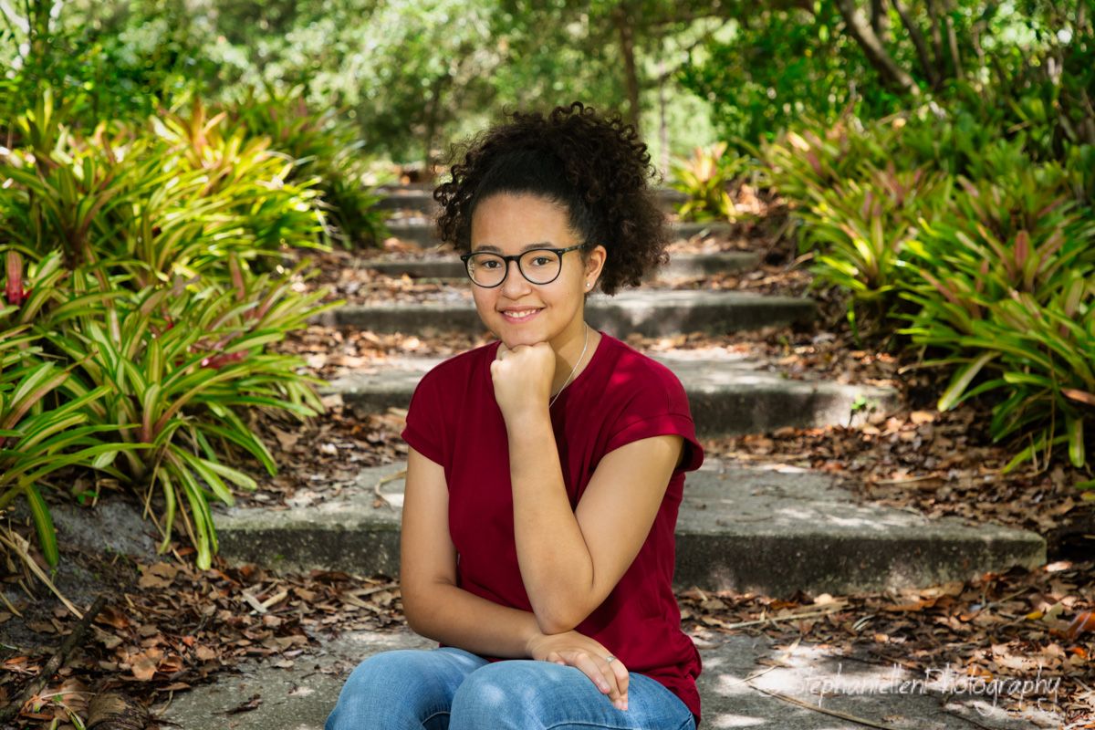 Teenage girl with curly hair and glasses smiling warmly while sitting on garden steps.