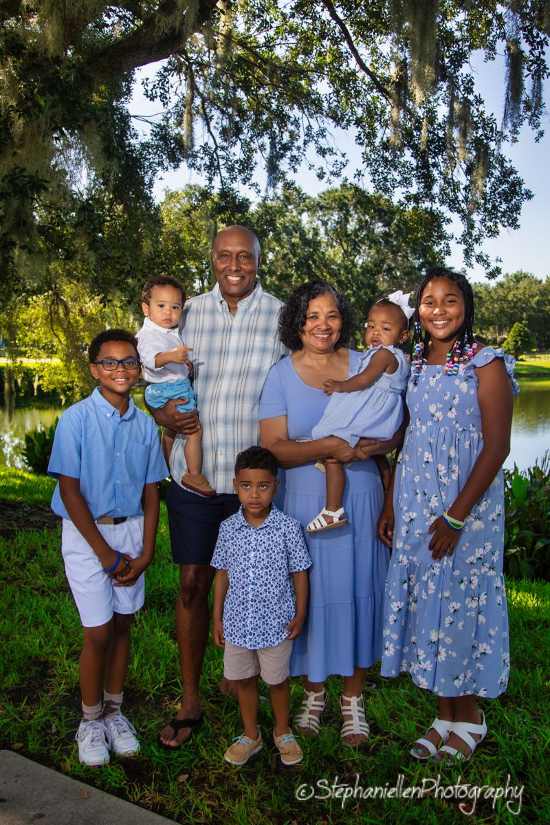 Multi-generational family of seven smiling together outdoors near a pond under a large oak tree in blue outfits.