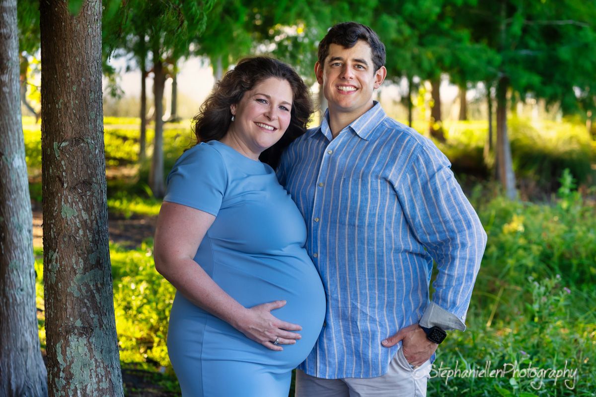 Happy pregnant couple posing outdoors in blue outfits among green trees during a maternity photography session.