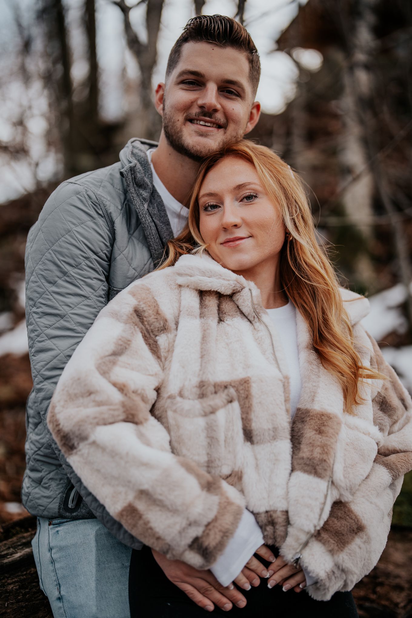 Smiling couple posing together in winter woods, woman wearing plaid fur coat.