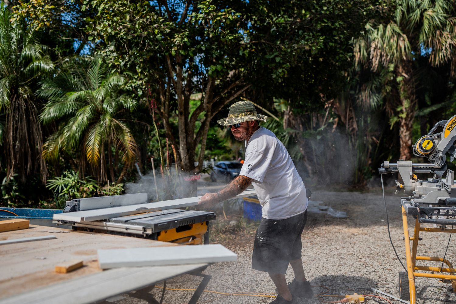 Worker cutting white boards with a table saw outdoors surrounded by tropical trees and sawdust.