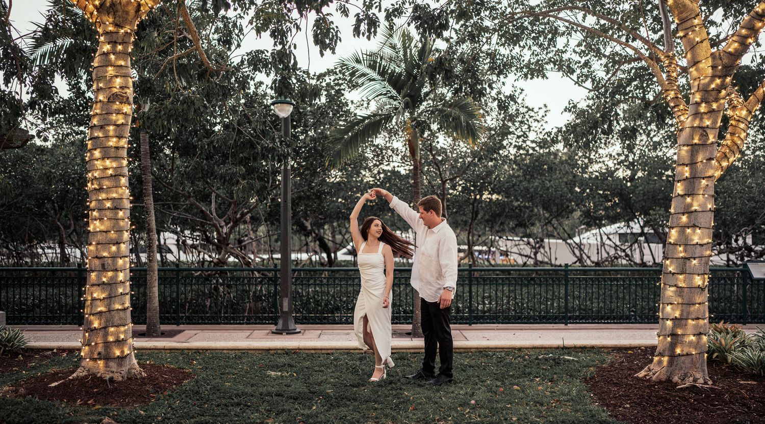 Happy couple dancing outdoors between fairy light-wrapped palm trees at twilight.