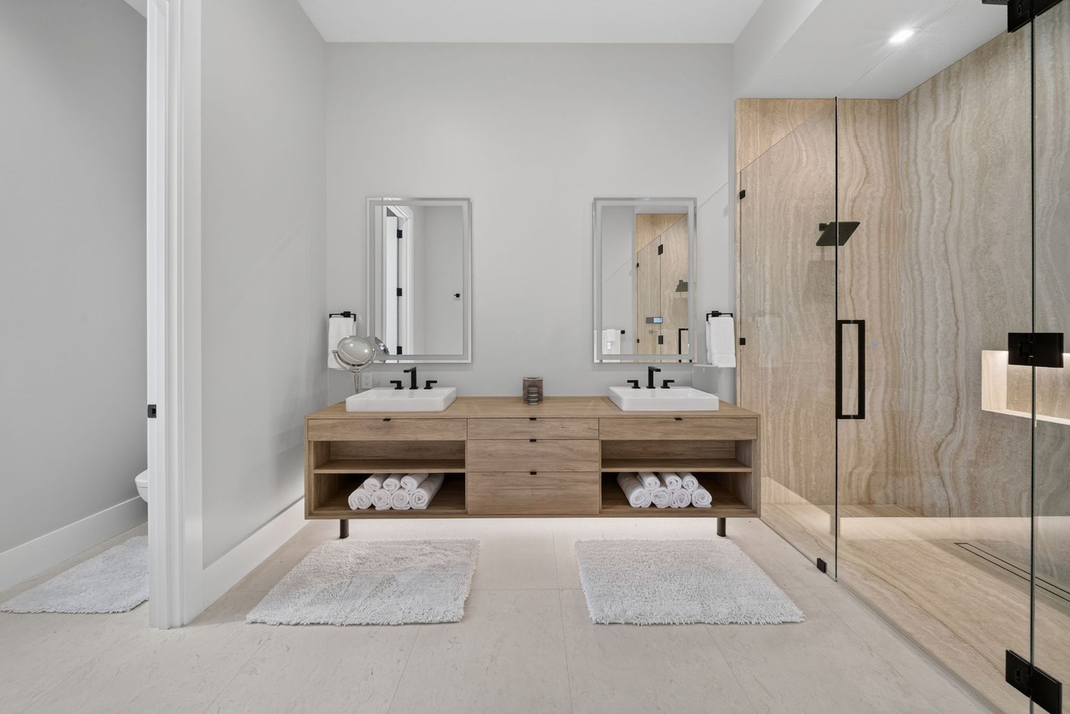 Modern bathroom with double vanity, wood cabinetry, dual mirrors, white towels, and glass-enclosed shower.