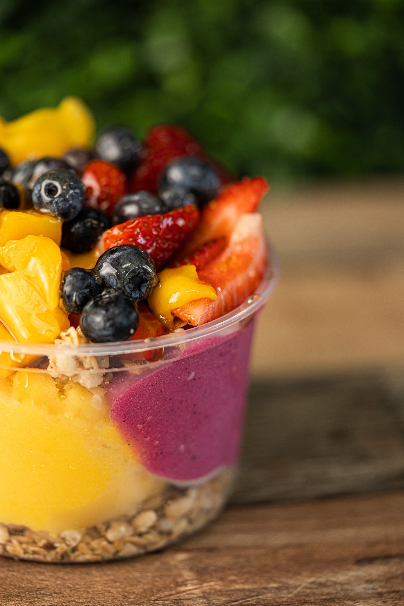 Fresh acai bowl with blueberries, strawberries, mango, and granola in plastic cup outdoors.