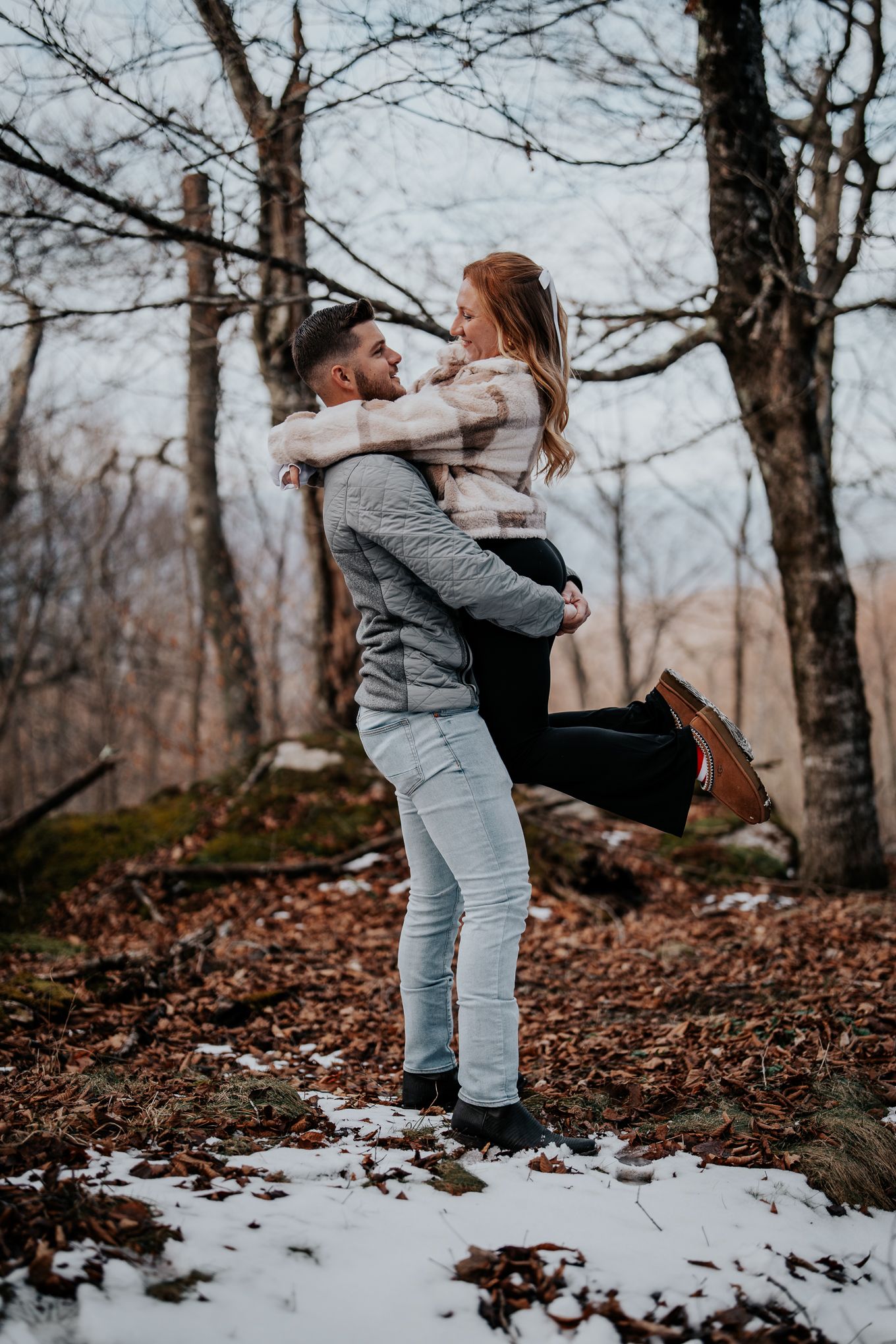 Couple embracing in snowy woodland setting during winter engagement photo session.