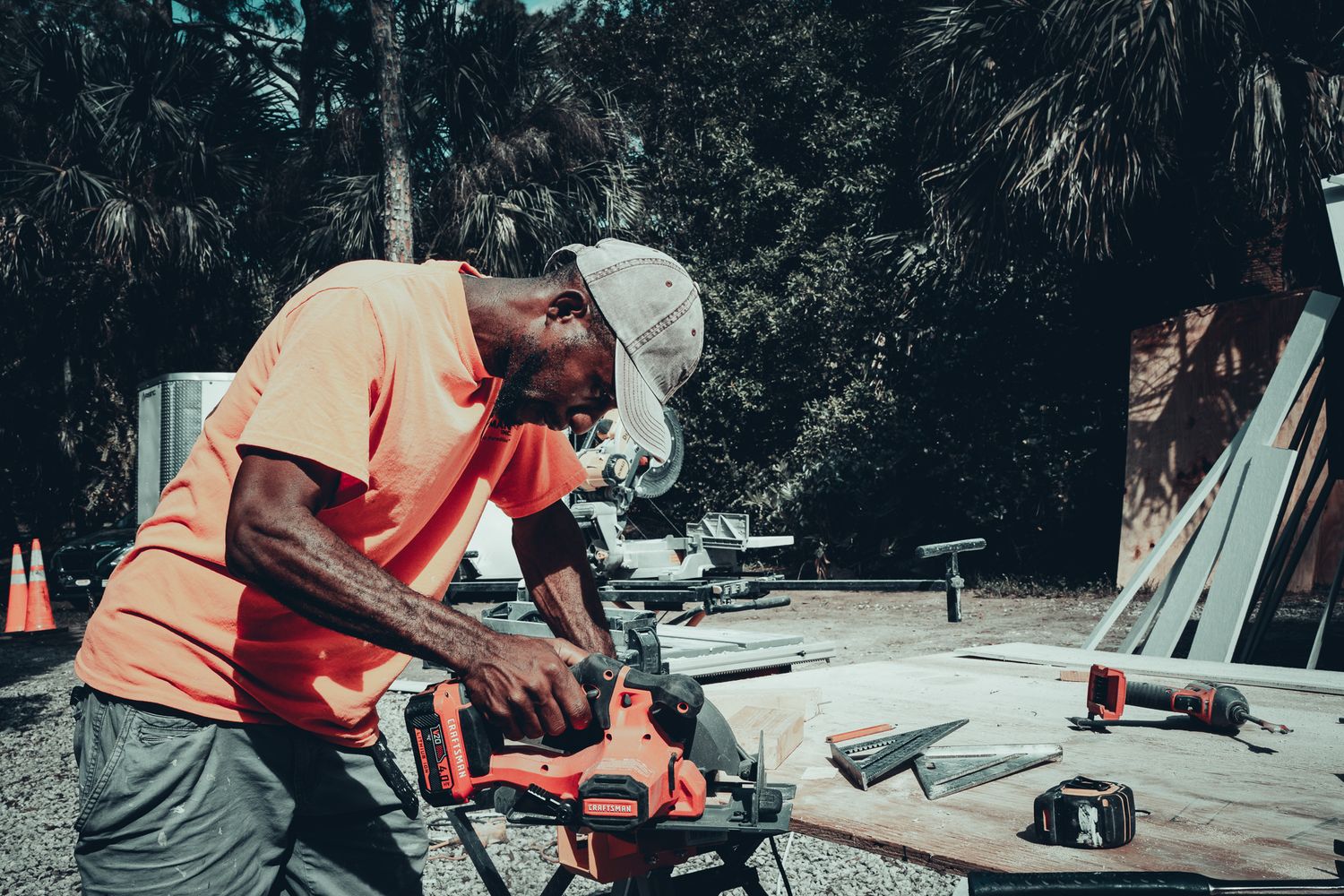 Construction worker in orange shirt operating red power saw cutting siding boards outdoors.