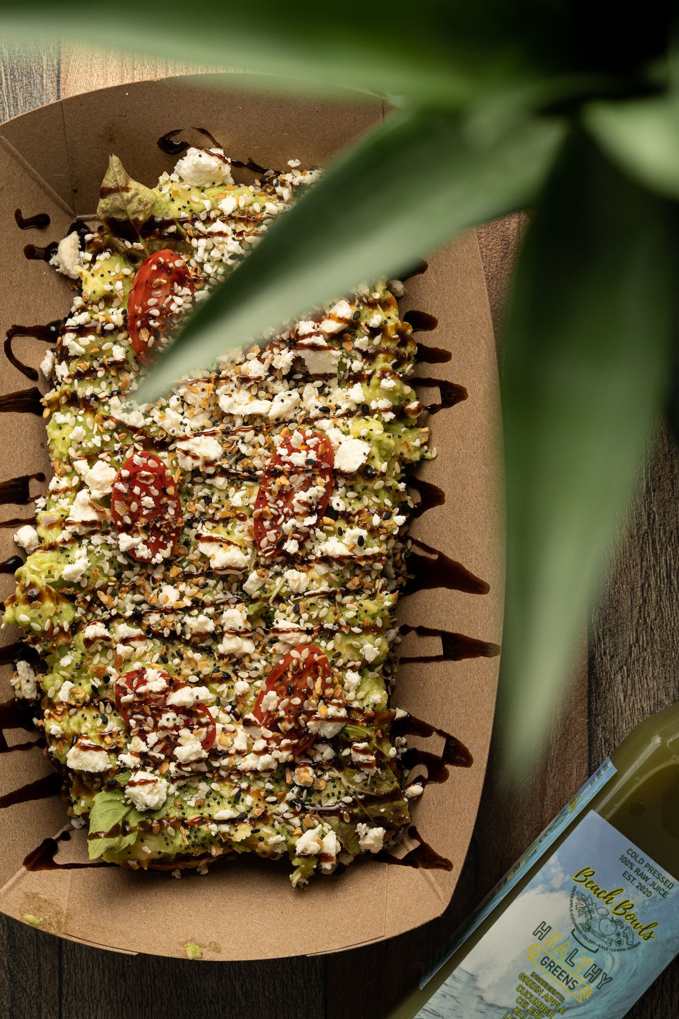 Gourmet flatbread topped with pesto, feta cheese, cherry tomatoes, and balsamic glaze served in a kraft paper tray.