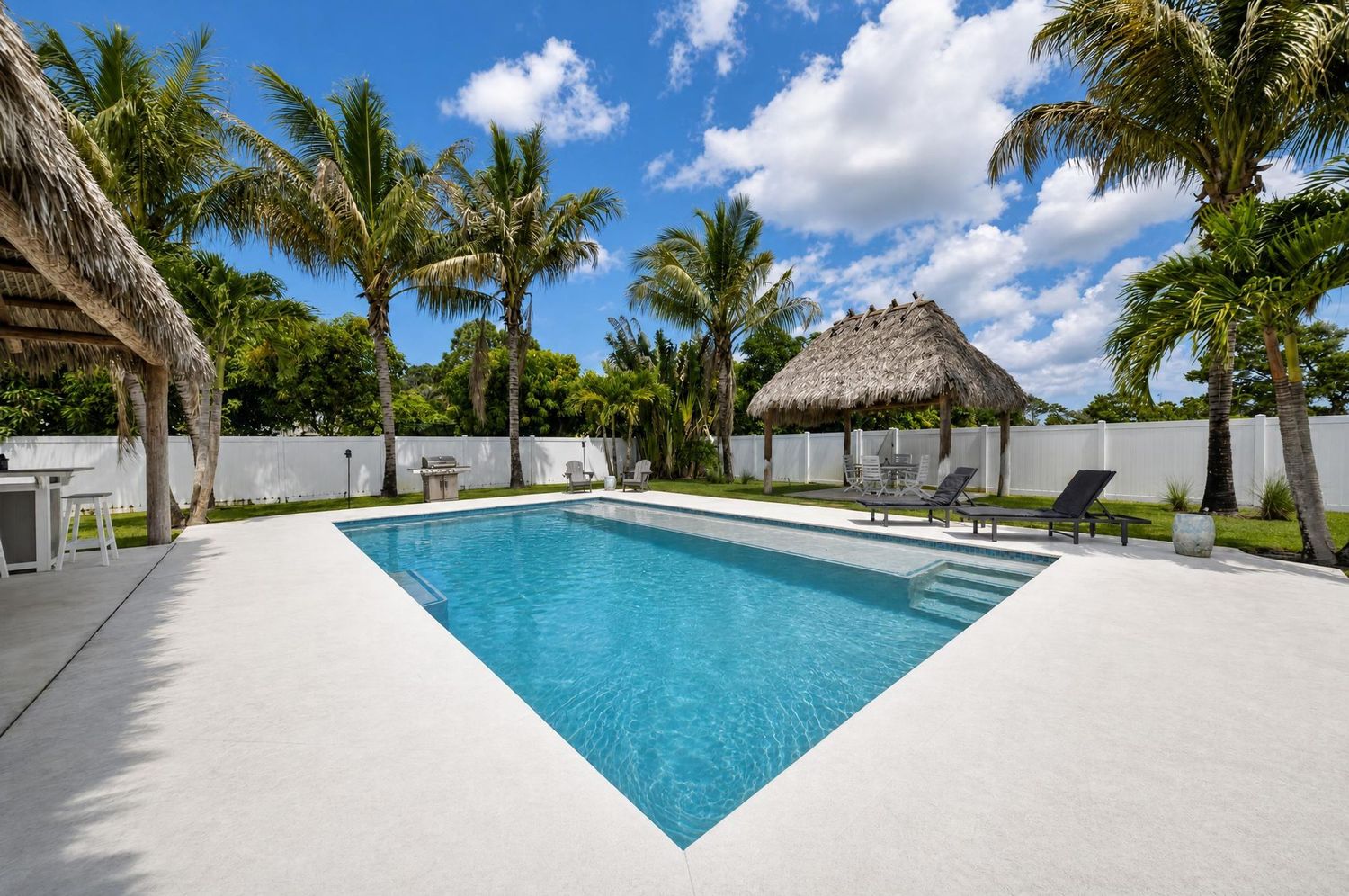 Tropical backyard oasis featuring rectangular pool, tiki hut, palm trees, and white privacy fence.