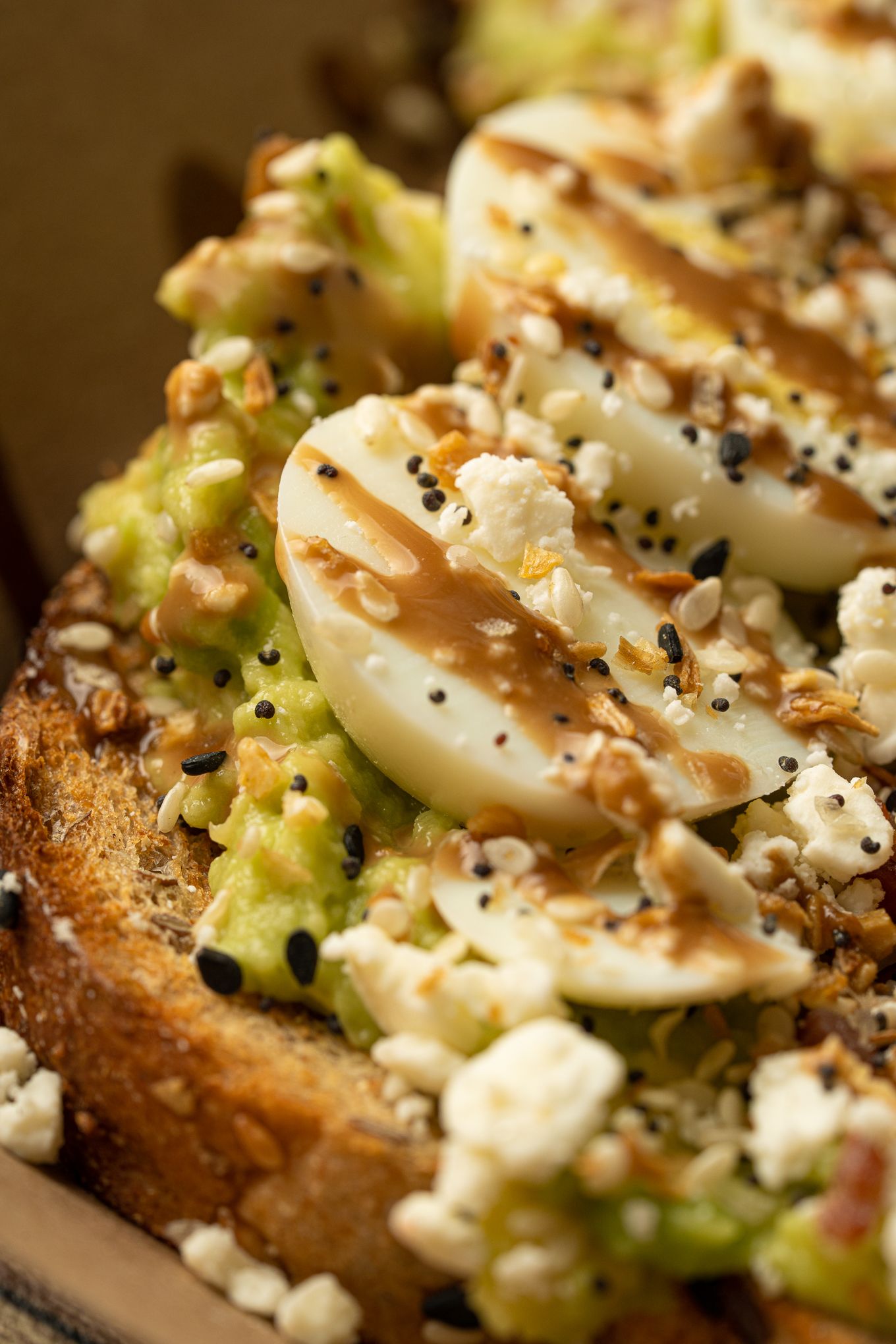 Close-up of avocado toast topped with sliced eggs, feta, seeds, and drizzled sauce on bread.