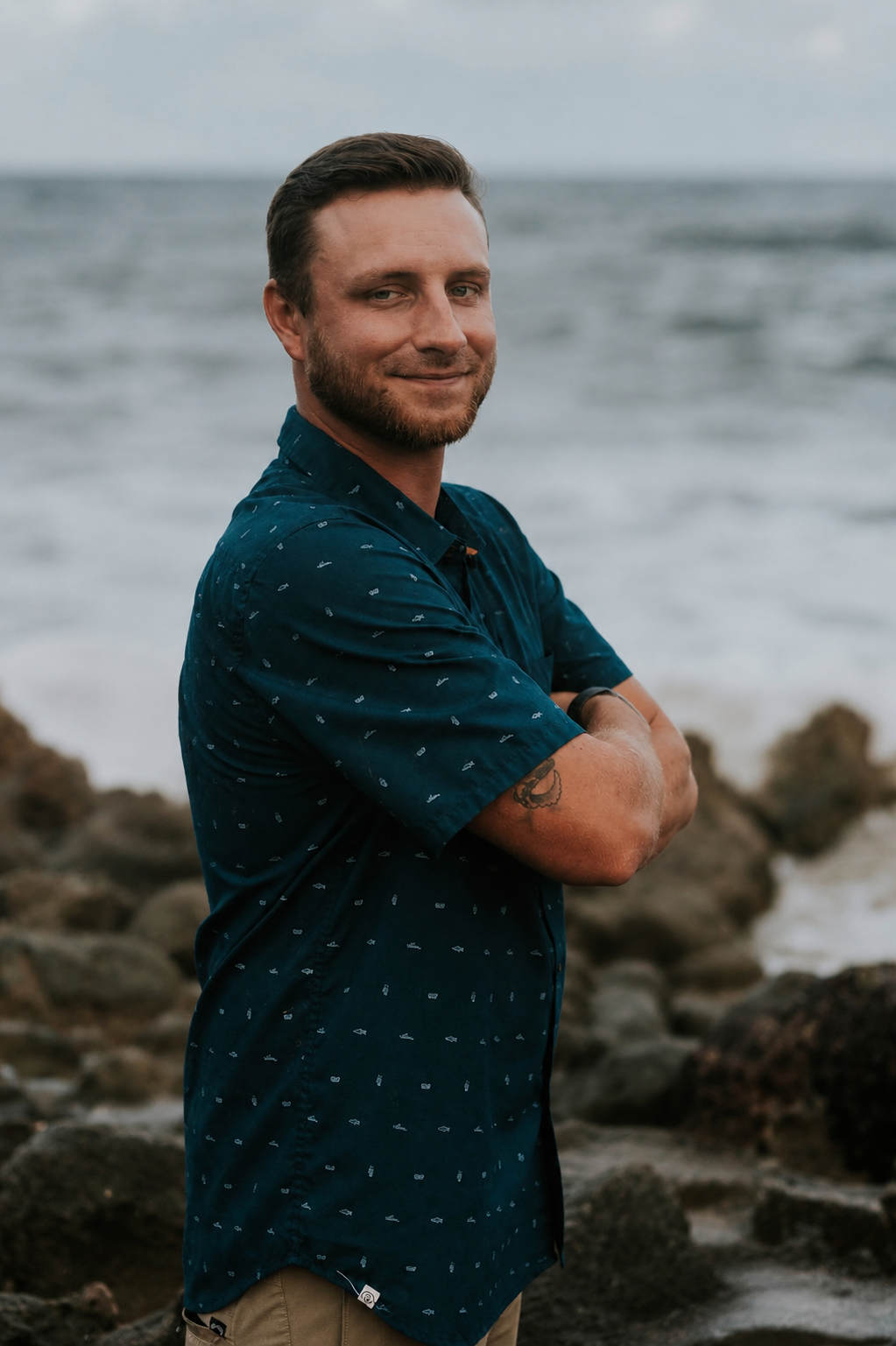 Smiling man with tattoo in teal shirt standing on rocky ocean shoreline with arms crossed.