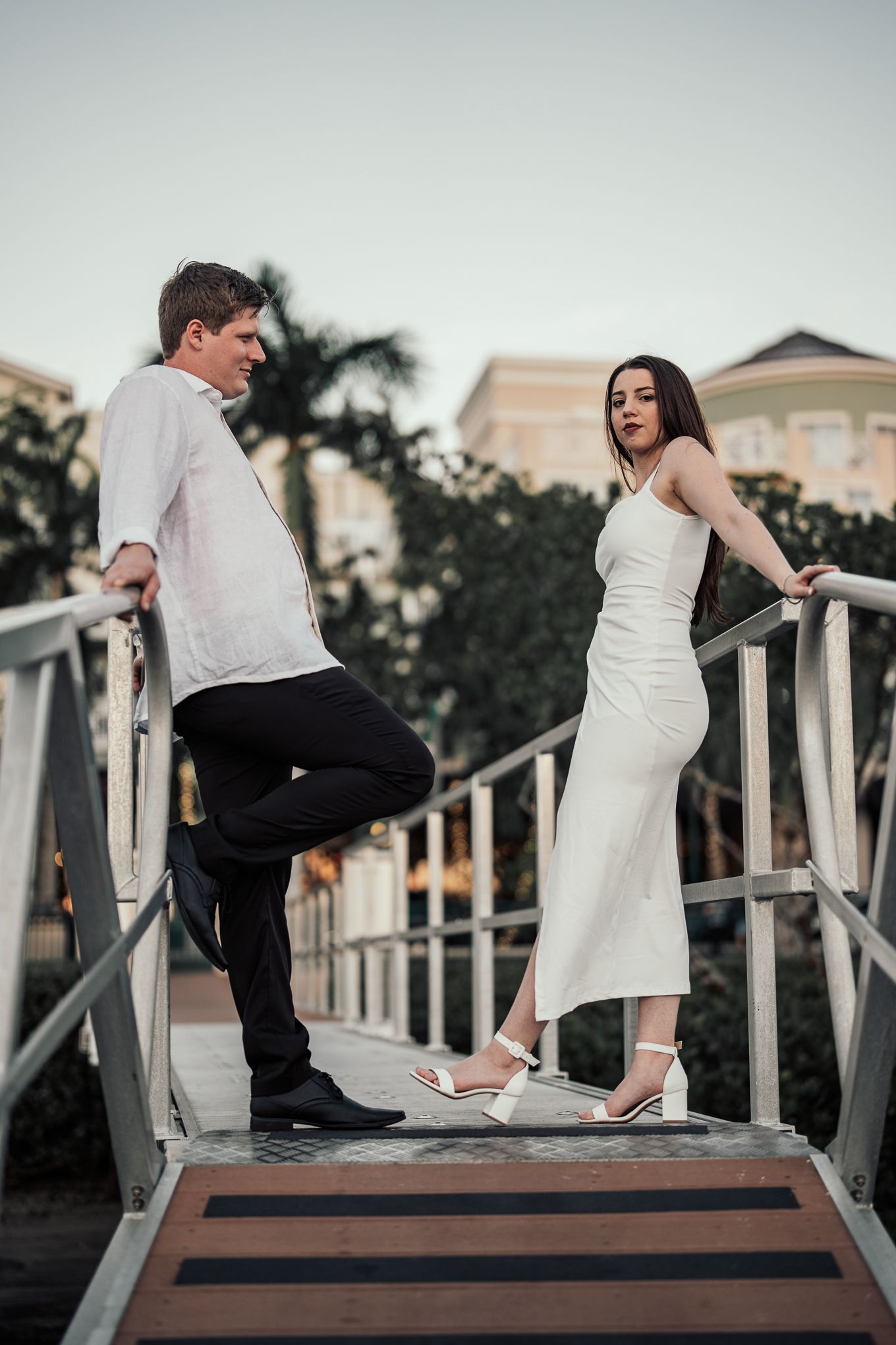 Stylish couple in white outfits posing on bridge ramp at tropical resort at sunset.
