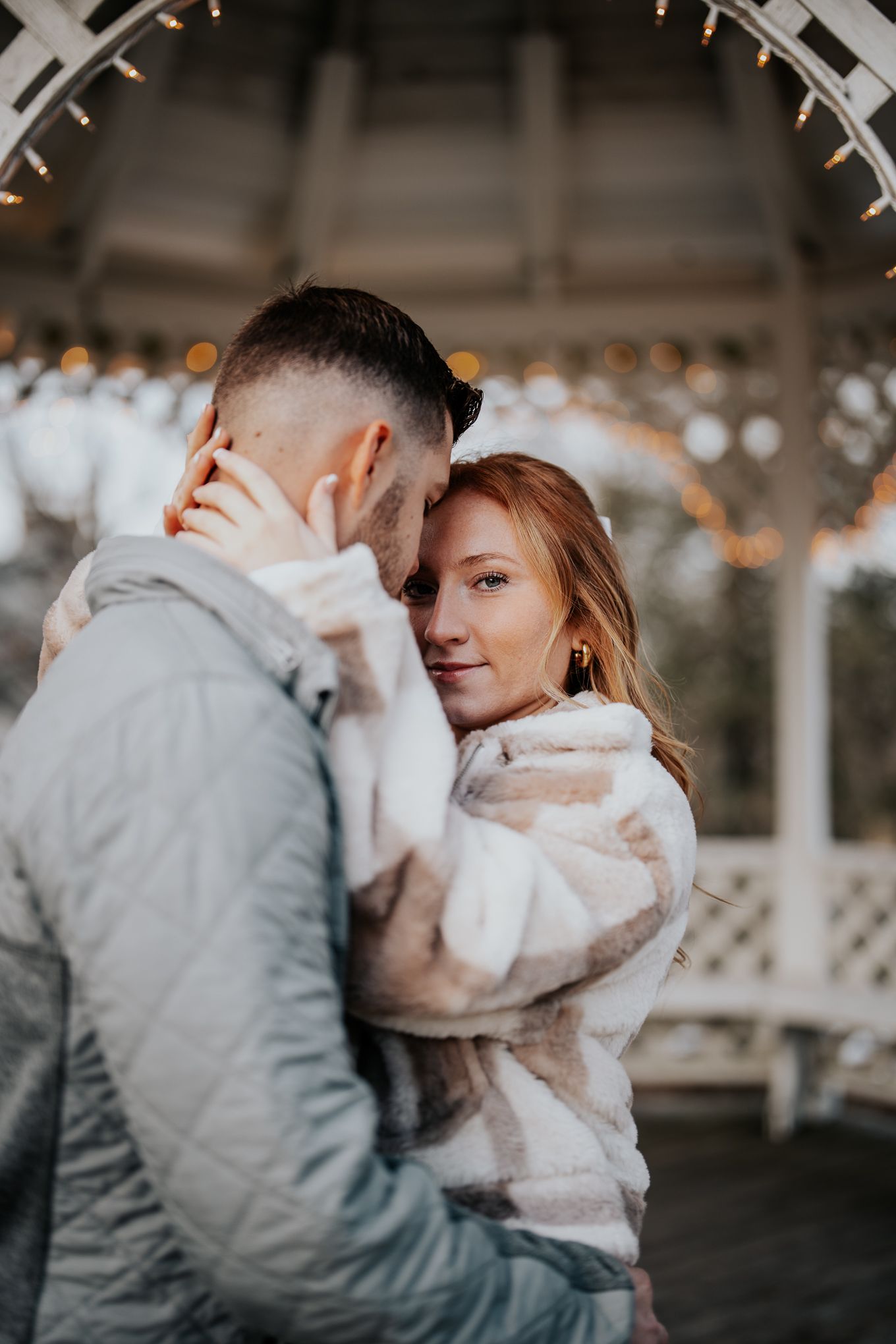 Romantic couple embracing near gazebo with string lights in winter setting.