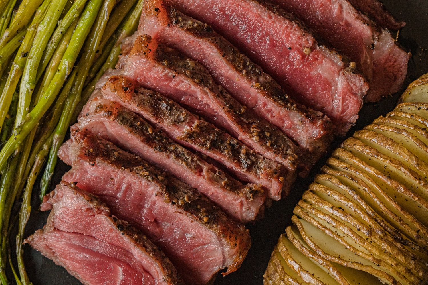 Close-up of perfectly sliced medium-rare steak with seasoned crust, served alongside asparagus and potato.